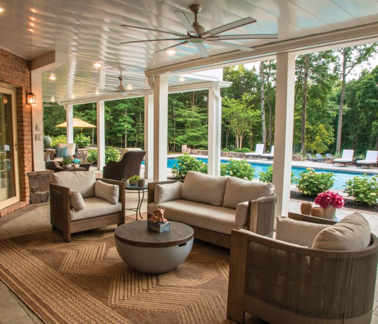 An outdoor room beneath a raised deck creates a seamless transition for indoor-outdoor entertaining. Beyond the covered area, a resort-style pool beckons.