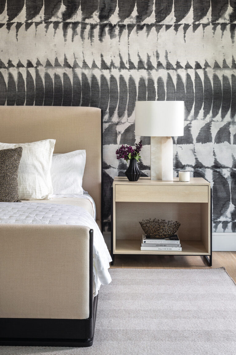 Graphic Holly Hunt wallpaper contrasts with the light wood of an Olivya Stone bedside table in a bedroom.