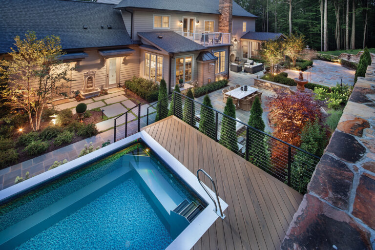 Seen from atop a high retaining wall, a multi-zone rear yard paved in bluestone features an elevated dipping pool, an outdoor kitchen and lounging and dining areas. Plantings delineate the zones.