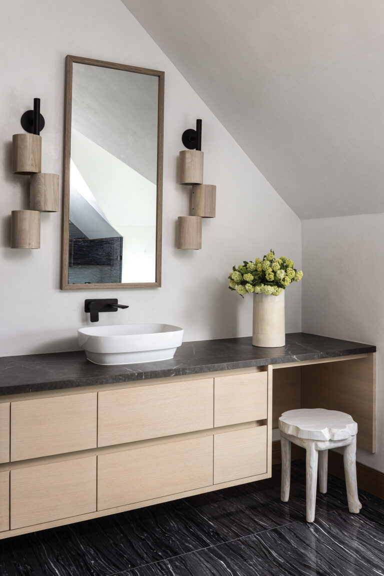 A soft, tonal mix of stone, wood and brushed metal completes a spa-like primary bath featuring eye-catching Allied Maker sconces.