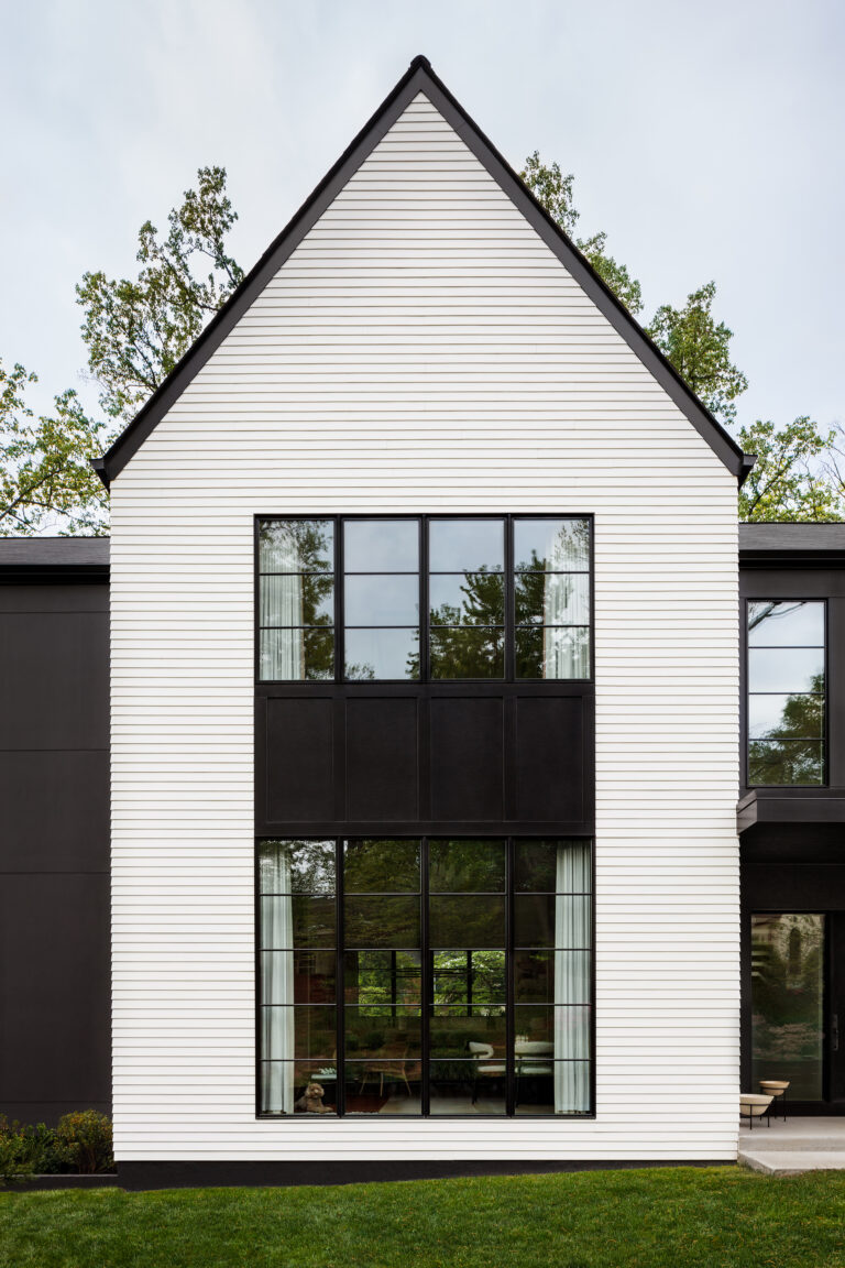 A steep gable and two-story steel-framed window wall dominate the facade, with white siding and black trim creating a high-contrast exterior.