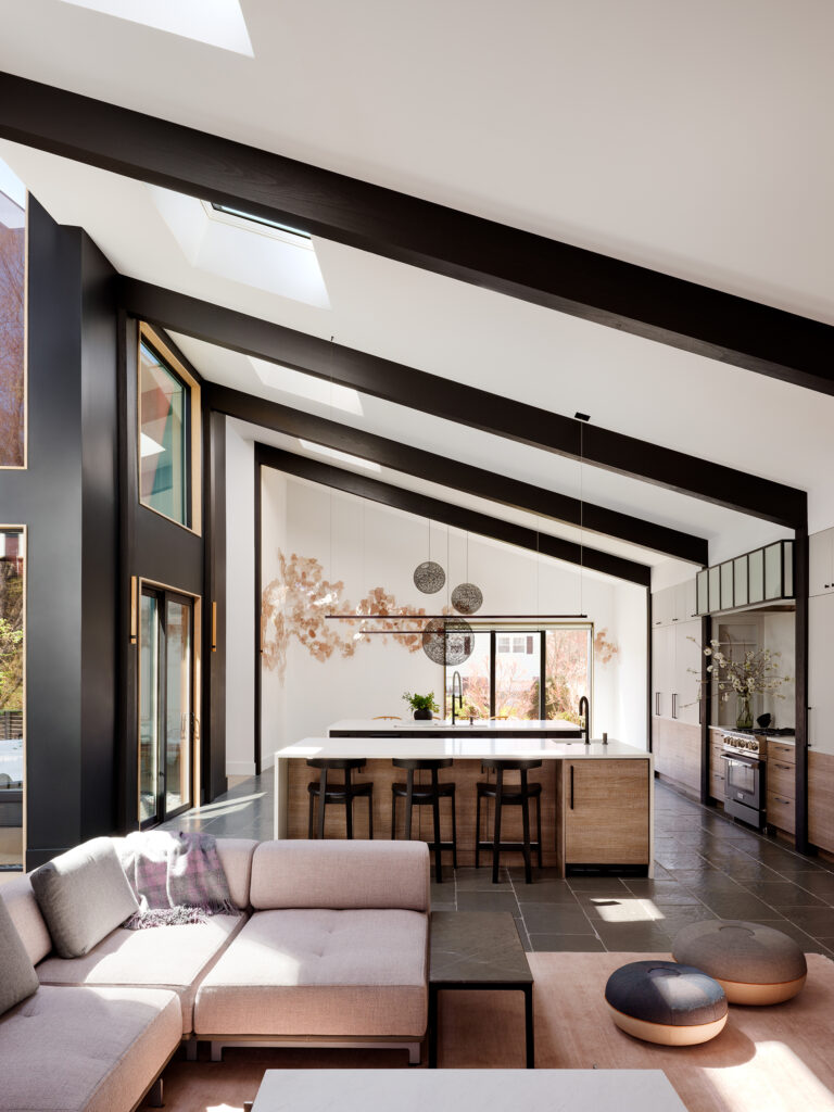 Dramatic raked ceilings and bold dark beams crown a double-height living space, where a kitchen island below grounds the room's soaring geometry.